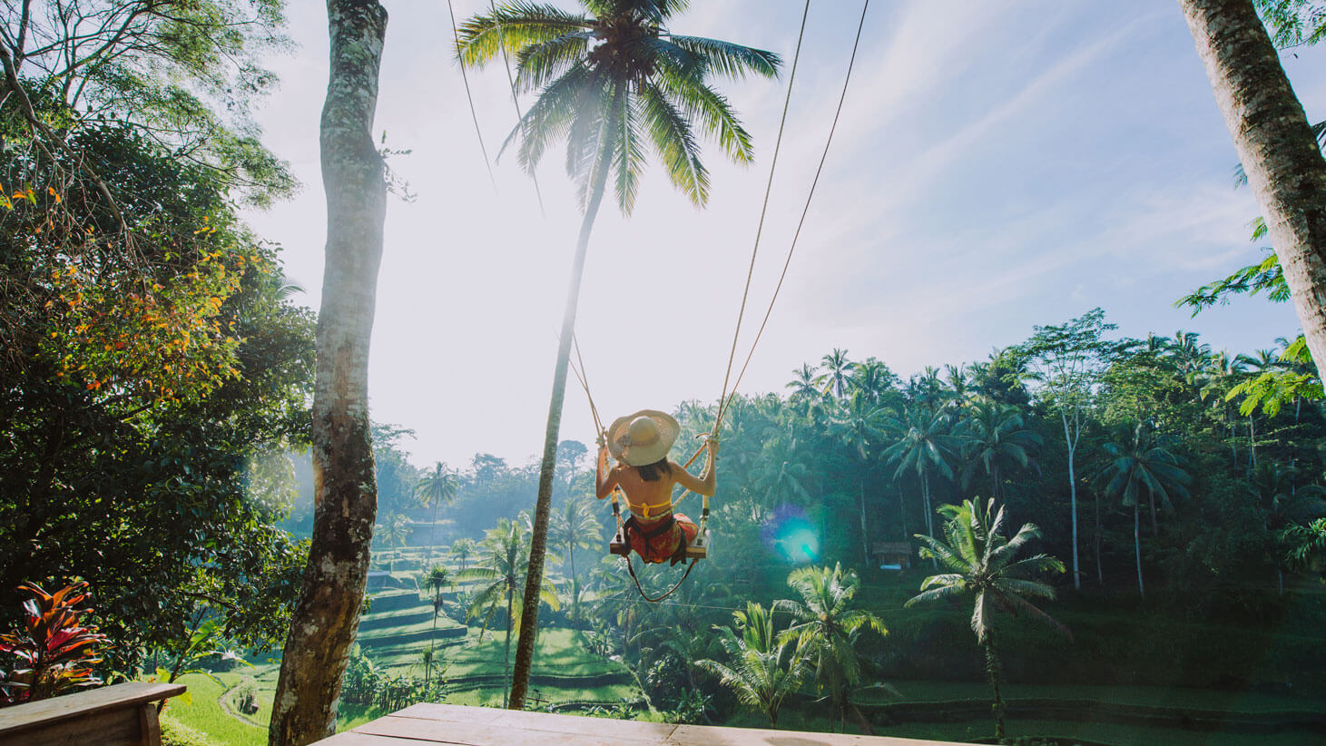 Glückliche Frau schaukelt im Urlaubsoutfit mit einer Schaukel unter Palmen, die Sonne scheint, der Himmel ist blau