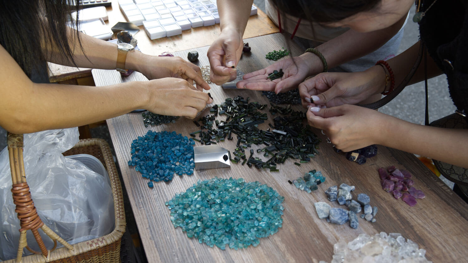 Edelsteinmarkt in Chanthaburi, Thailand: Man sieht drei Frauen die sich kleine Häufchen ungeschliffener Steine ansehen, die auf einem braunen Tisch liegen
