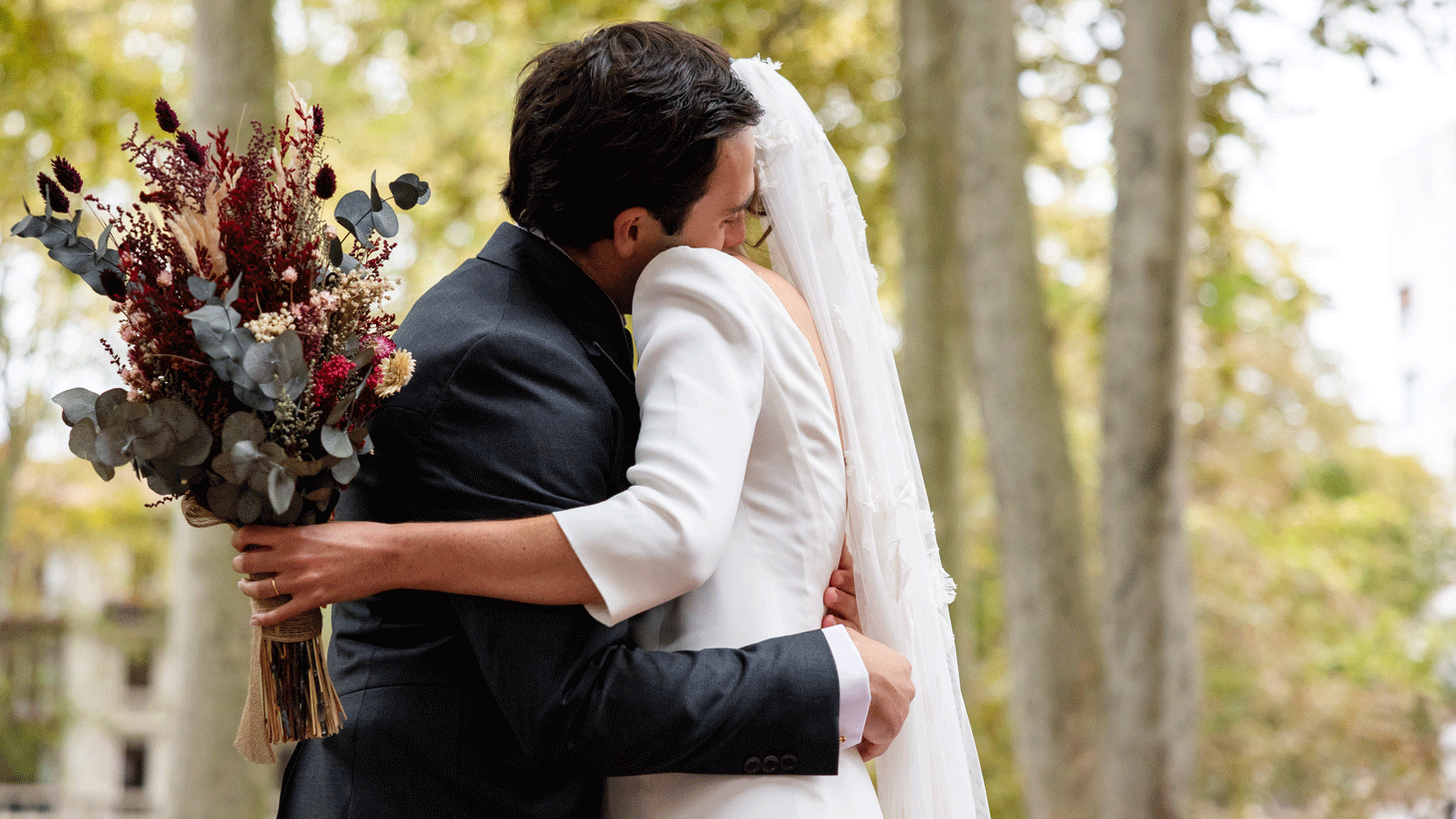 Hochzeitspaar umarmt sich in einem Outdoor Setting. Er trägt einen schwarzen Anzug, sie ein weißes Kleid und hält einen Blumenstraß in der Hand. Die Gesichter des Paares sind verdeckt.